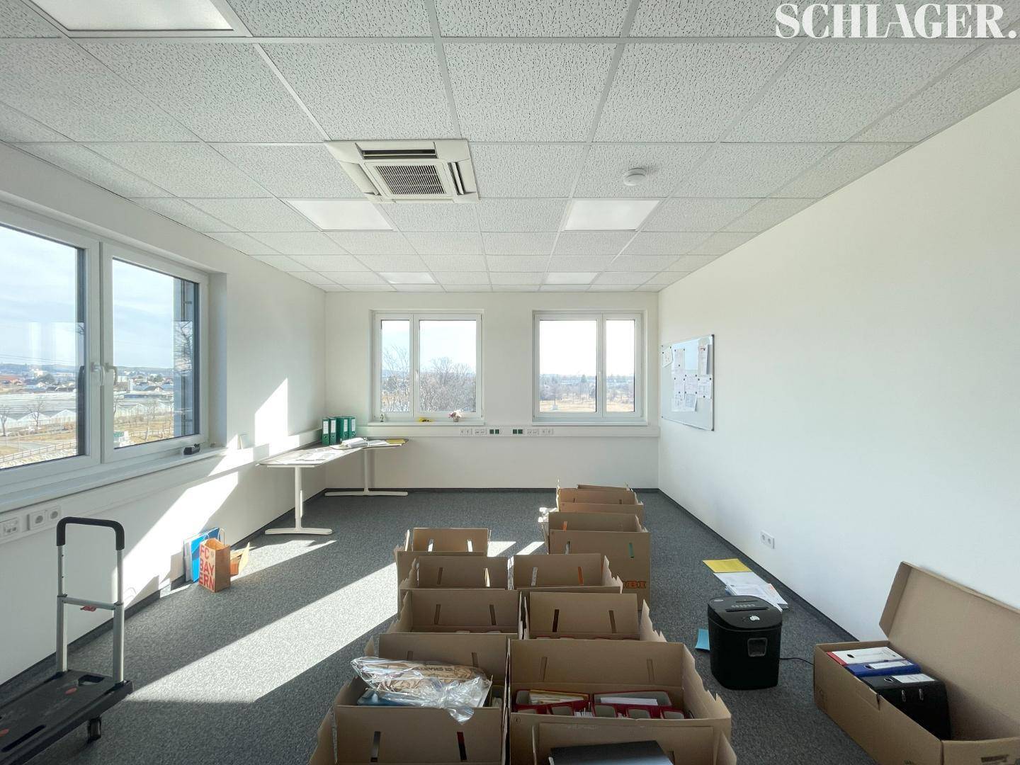 Helles Büro mit Teppichboden, Fenstern und mehreren Umzugskartons auf dem Boden, bereit für den Umzug.