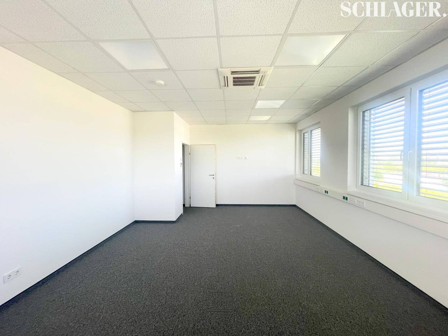 Geräumiges, leeres Büro mit Teppichboden, weißen Wänden und einer Tür, die zu einem weiteren Bereich führt.