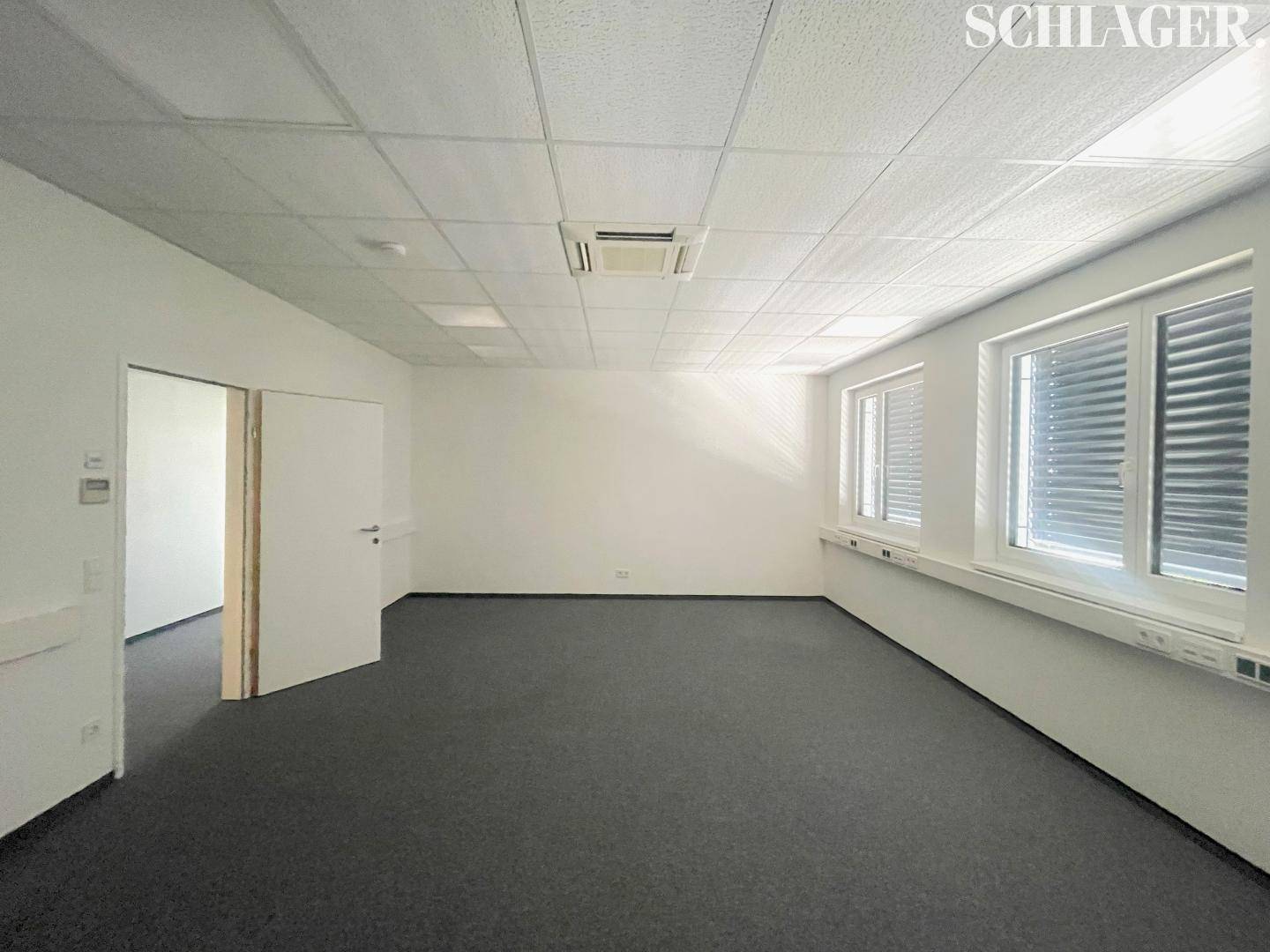Leeres Büro mit Teppichboden, weißen Wänden und einer offenen Tür, die den Zugang zu einem Nebenraum ermöglicht.