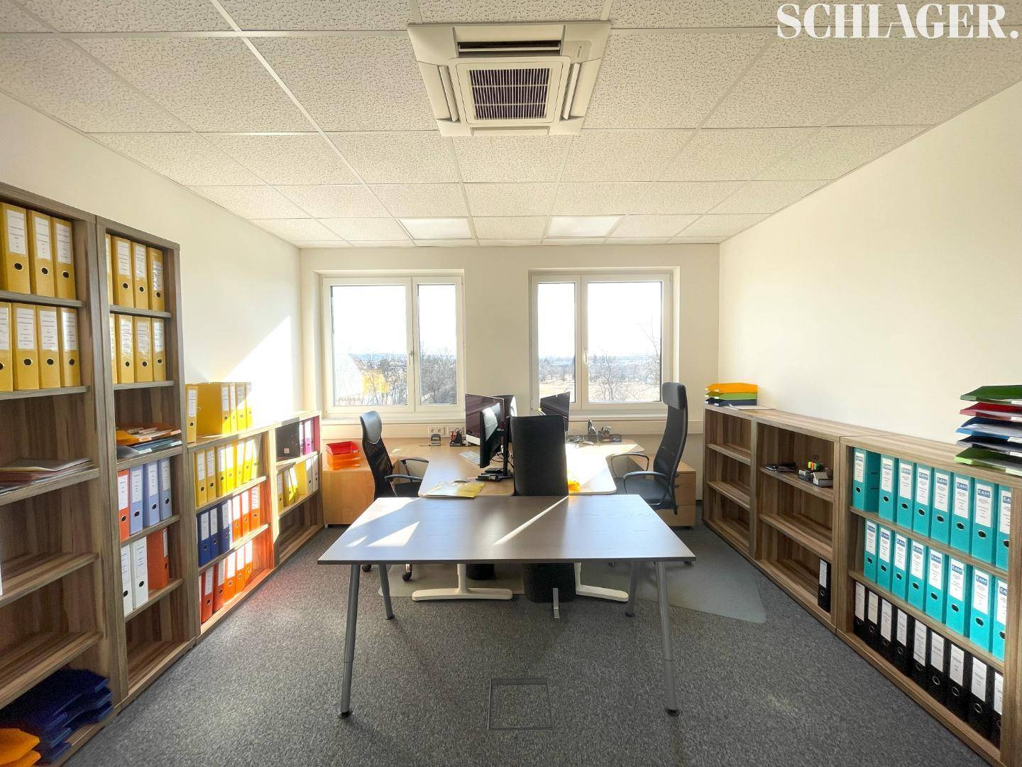 Möbliertes Büro mit mehreren Arbeitsplätzen, Schreibtischen, Bürostühlen und Regalen voller Aktenordner.