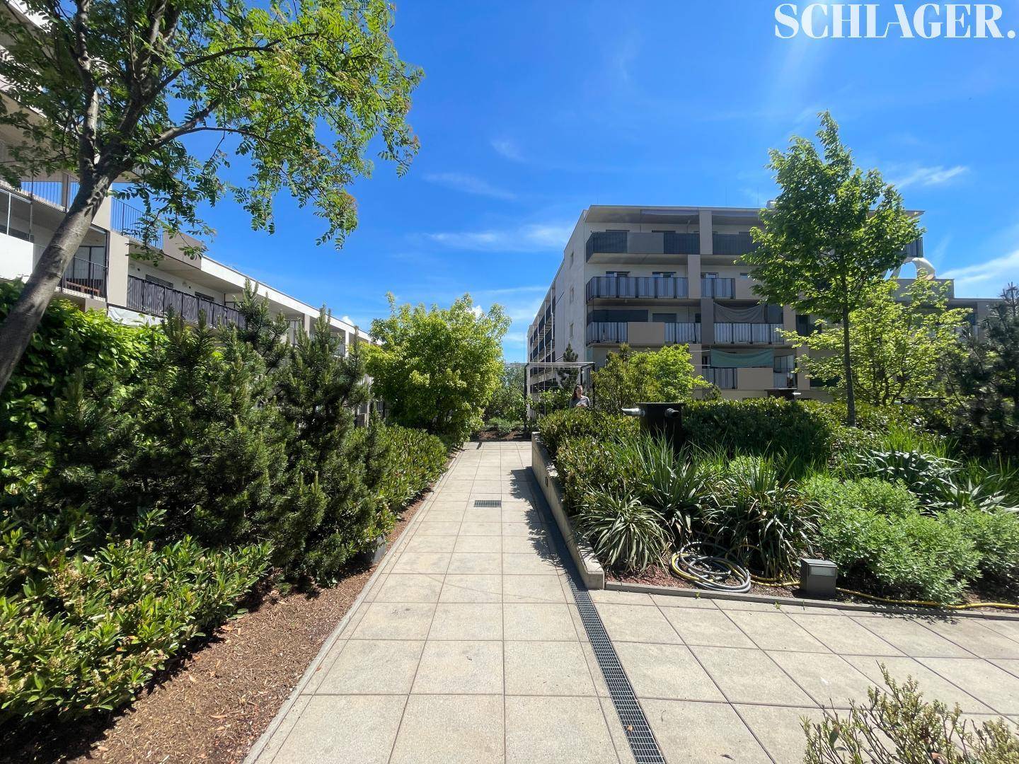 Gepflegter Gartenweg mit üppiger Bepflanzung, der zu den modernen Wohngebäuden im Hintergrund führt.