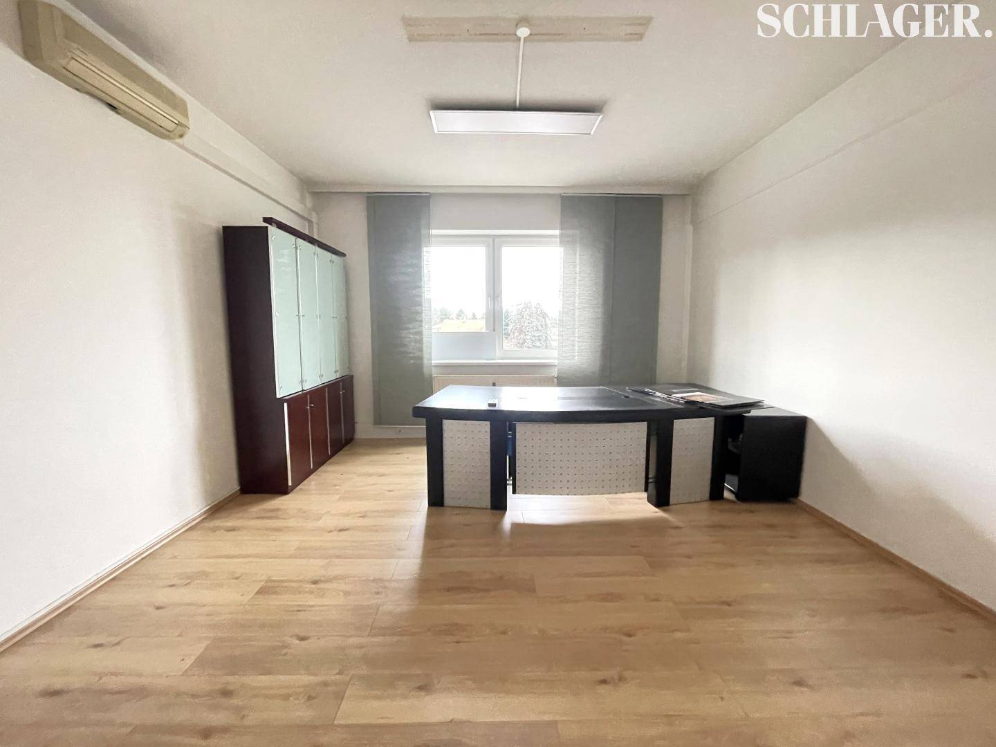 Helles Büro mit Holzboden, Schreibtisch und Schrank, gut beleuchtet durch ein großes Fenster.