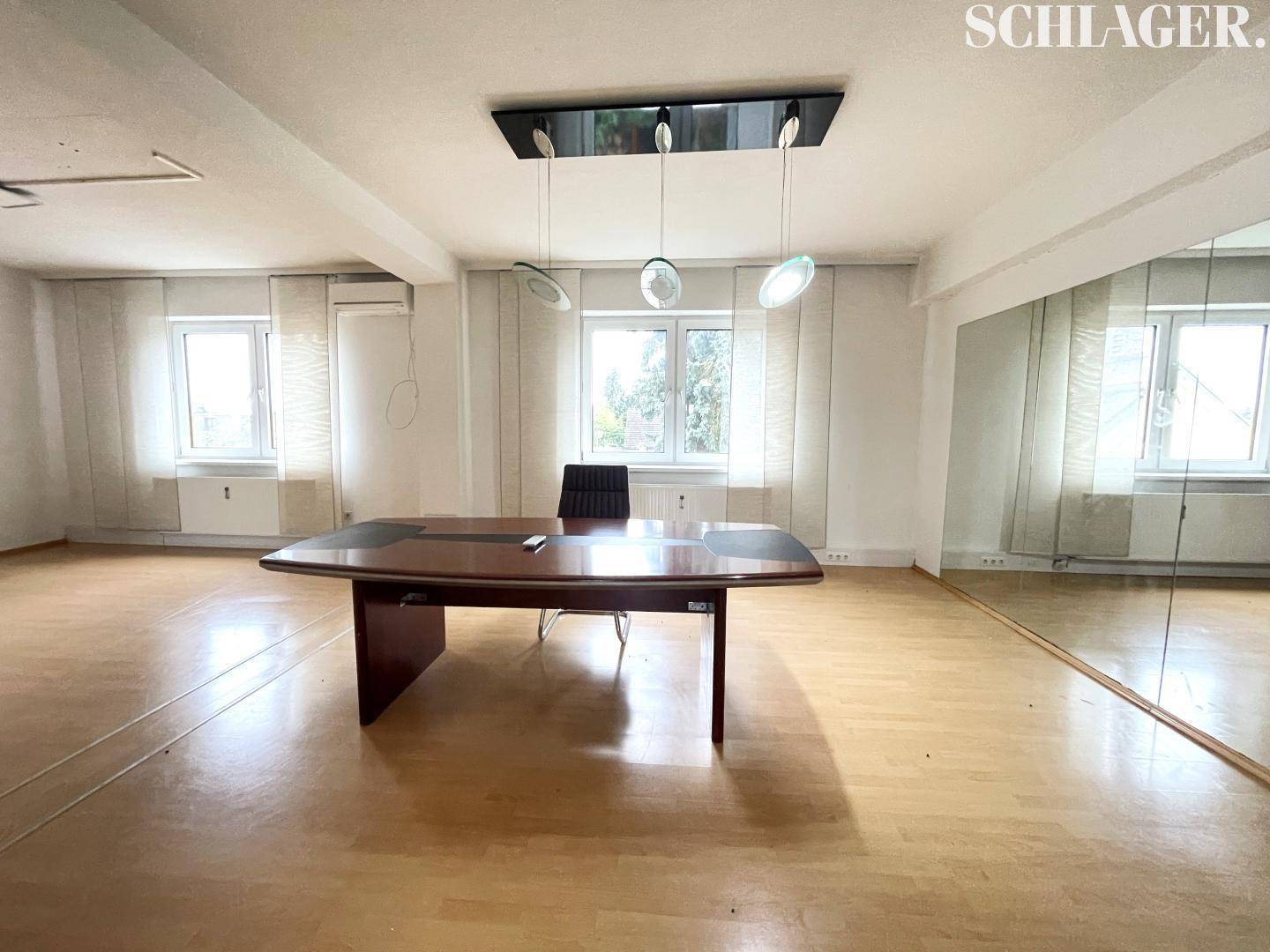 Geräumiges Büro mit Holzboden, großem Konferenztisch und Spiegelwand, gut beleuchtet durch Fenster.