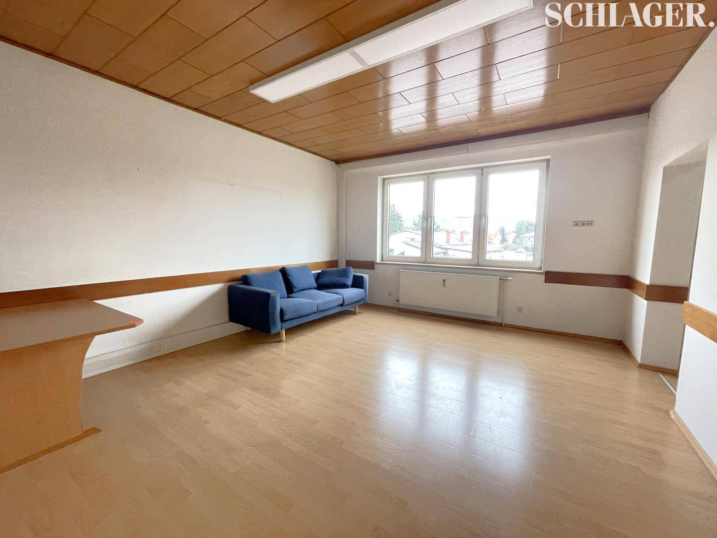 Heller Raum mit Holzboden, blauer Couch und Holzdecke, gut beleuchtet durch große Fenster.