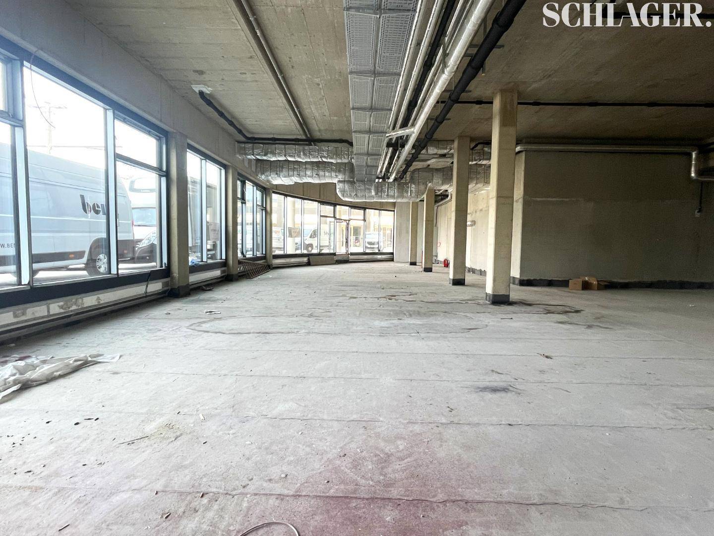 Langer, leerer Gewerberaum mit Betonboden, sichtbaren Installationen an der Decke und einer gebogenen Fensterfront.