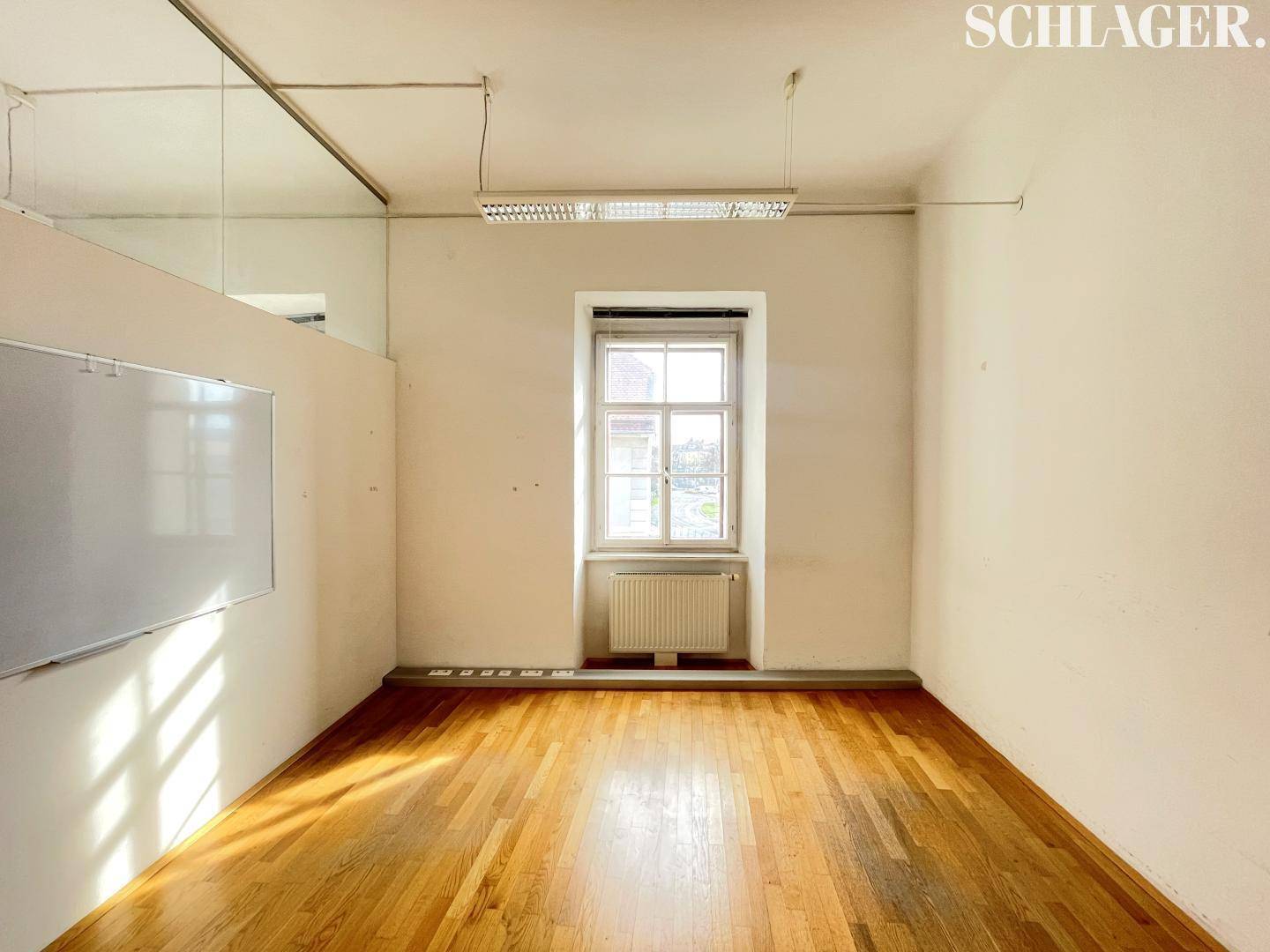 Kleiner Büroraum mit Holzboden, einem Fenster und einer weißen Tafel an der Wand.