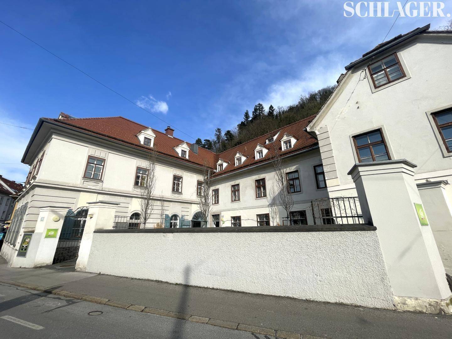 Ansicht eines historischen Gebäudekomplexes mit Innenhof und rotem Ziegeldach unter blauem Himmel.