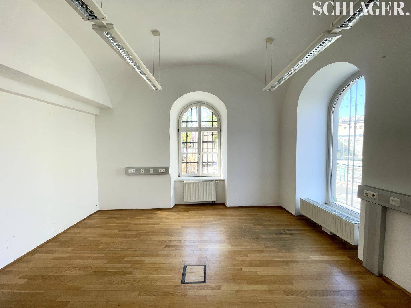 Geräumiger Büroraum mit Holzboden, gewölbter Decke und großen Fenstern, die viel Licht hereinlassen.