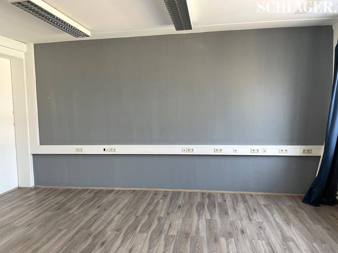 Graue Wand in einem Büroraum mit einer langen Reihe von Steckdosen und Laminatboden.