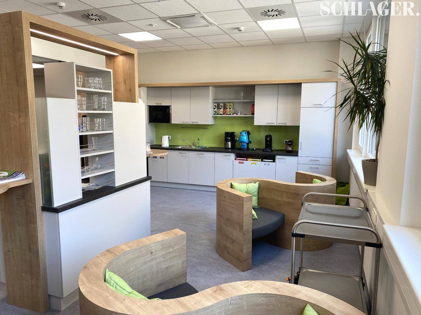 Moderne Büroküche mit hellen Schränken, grüner Rückwand und geschwungenen Holzsitzgelegenheiten.