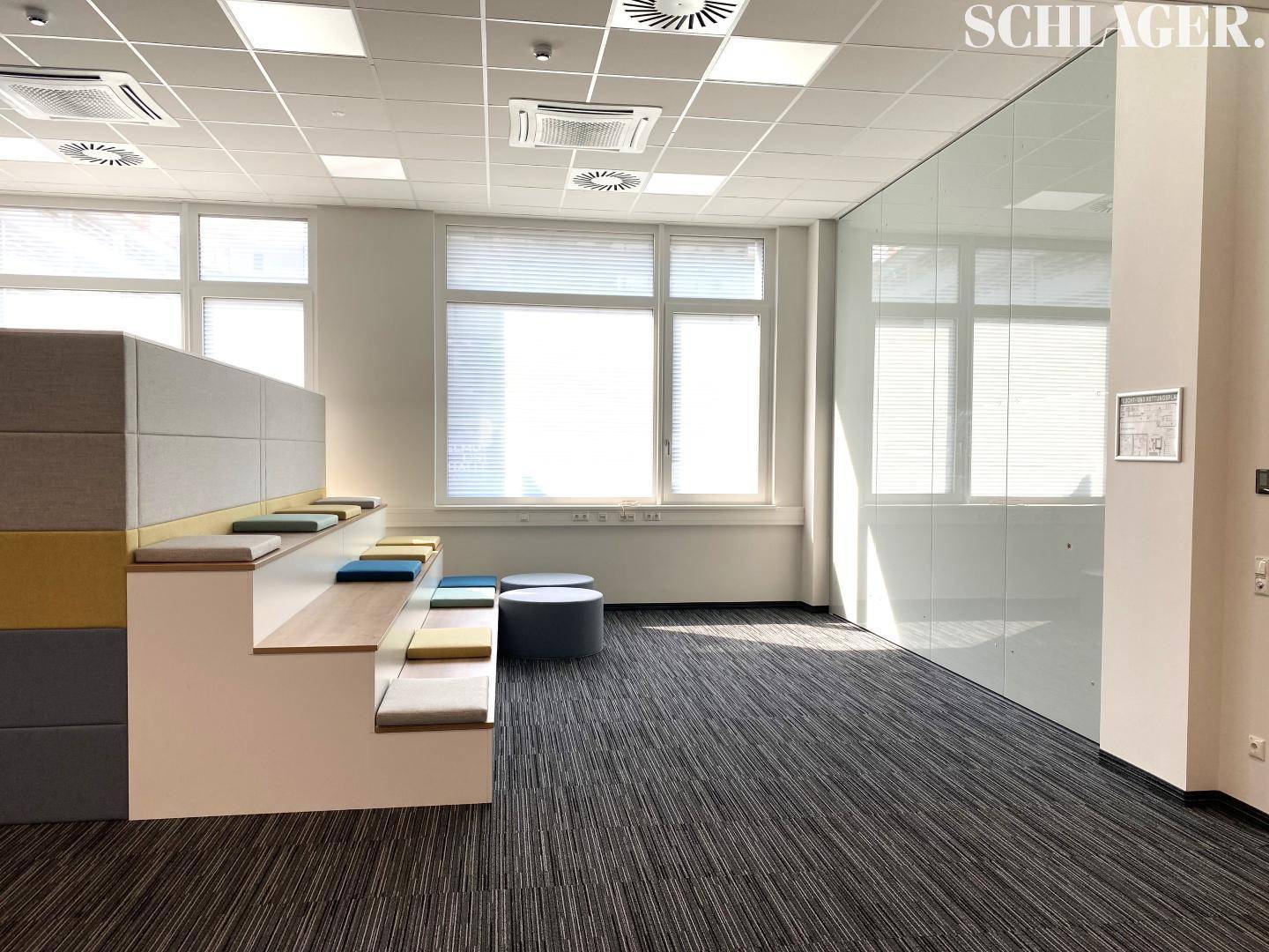 Heller Loungebereich im Büro mit gestuften Sitzgelegenheiten, großen Fenstern und viel Tageslicht.