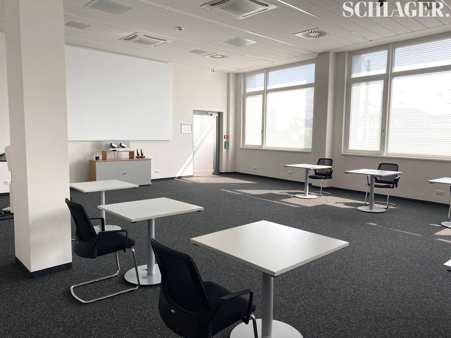 Heller Konferenzraum mit Teppichboden, großen Fenstern und flexiblen Tischen für Besprechungen.