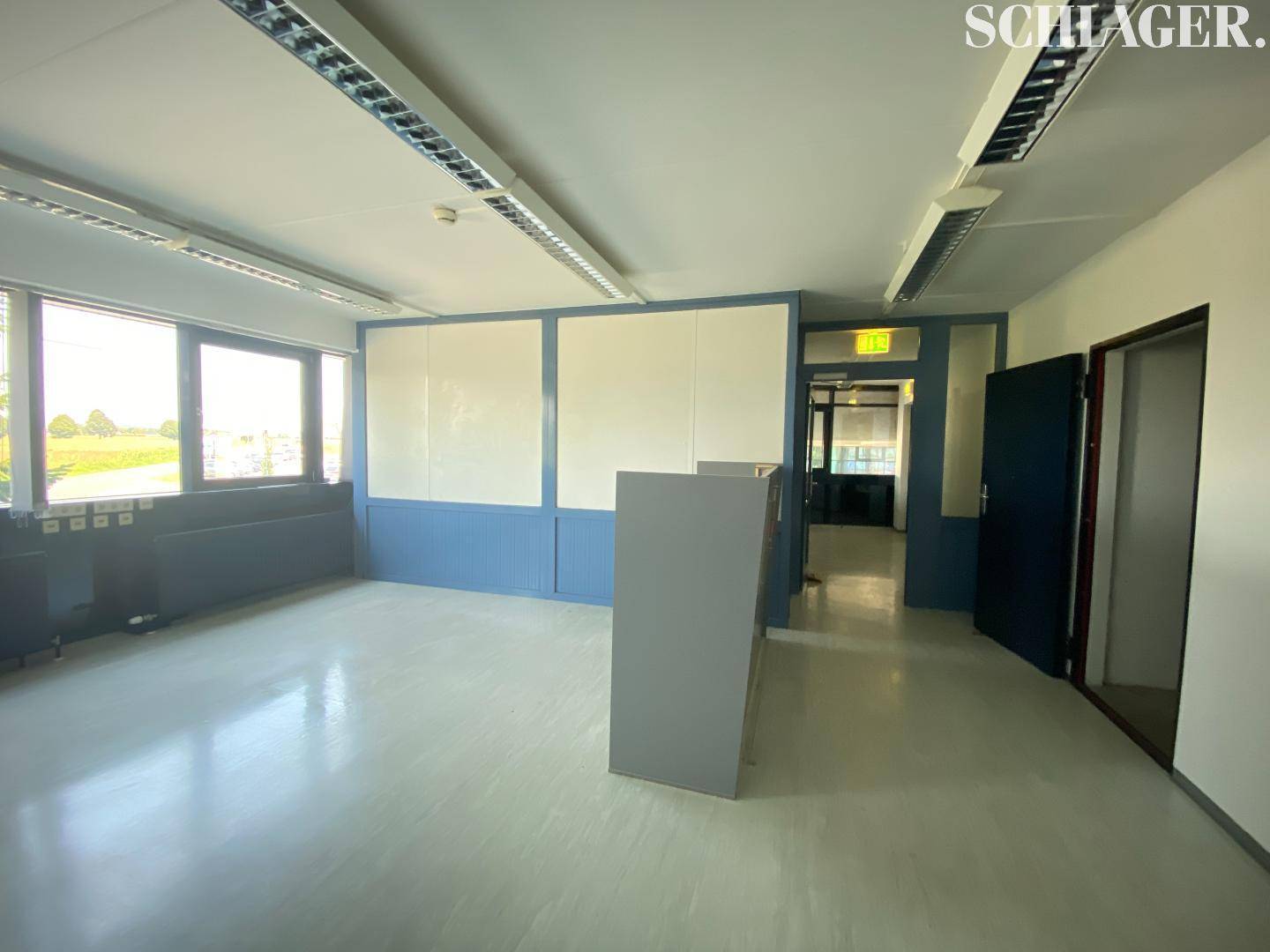 Leerer Bürobereich mit großen Fenstern, blauen Wänden und Zugang zu weiteren Räumen.