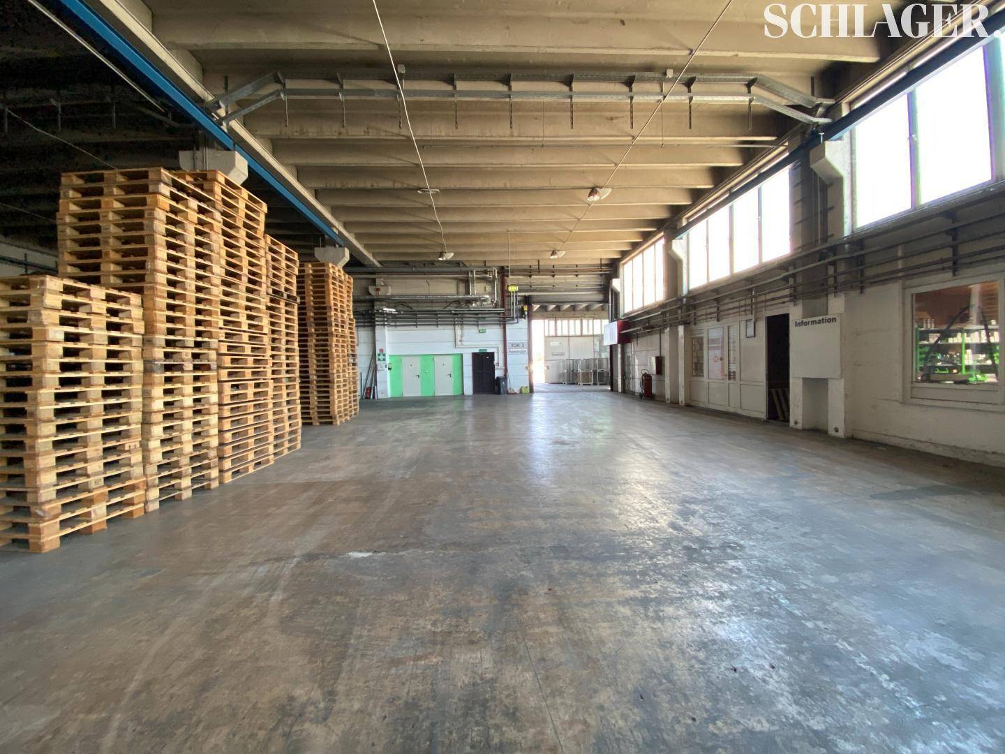 Weitläufige Lagerhalle mit gestapelten Paletten, hohen Decken und großen Fenstern, die viel Tageslicht hereinlassen.