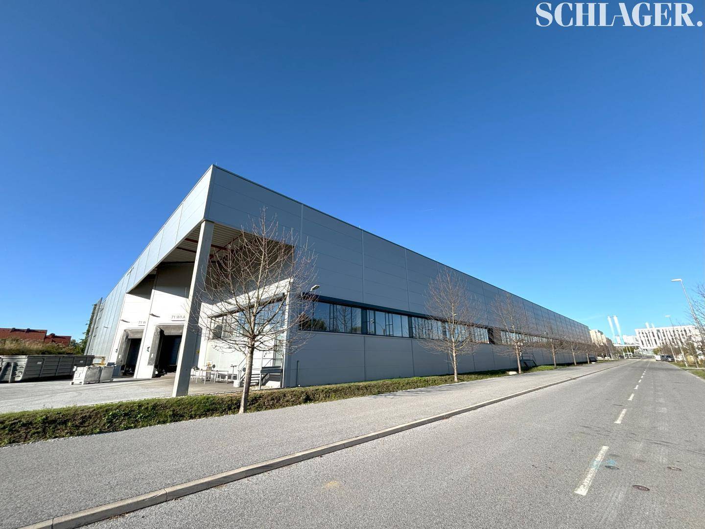Moderne Industriefassade mit großen Fenstern und Zufahrtsstraße unter klarem blauem Himmel.