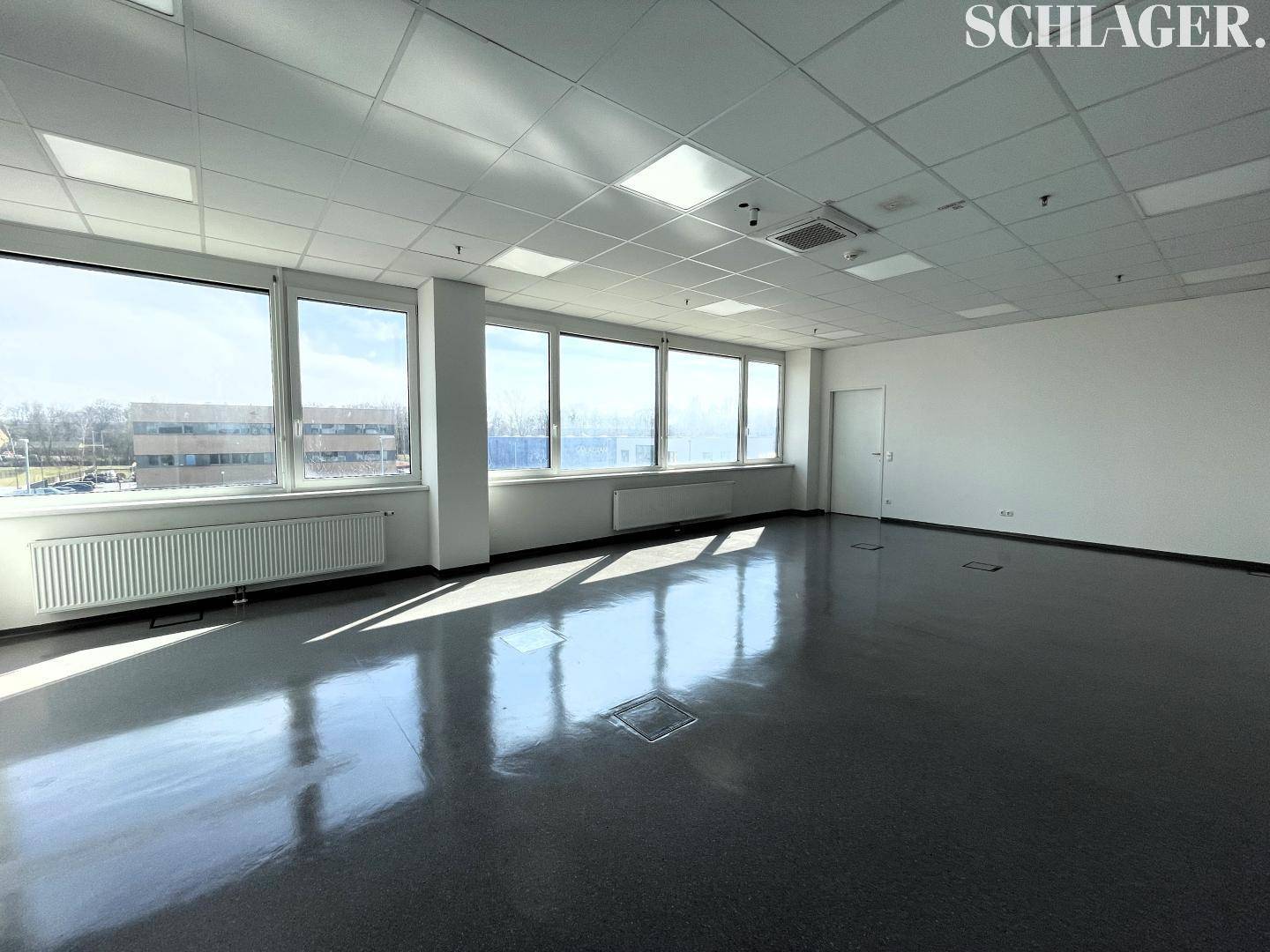 Helles, leeres Büro mit großen Fenstern, abgehängter Decke und Blick nach draußen.