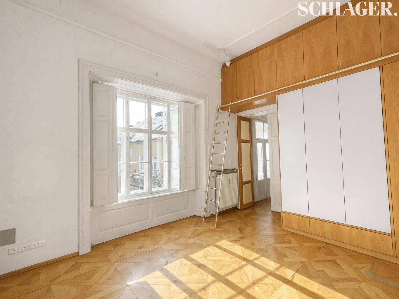 Heller Raum mit Fischgrätparkett, Einbauschränken und einem großen Fenster mit Fensterläden.