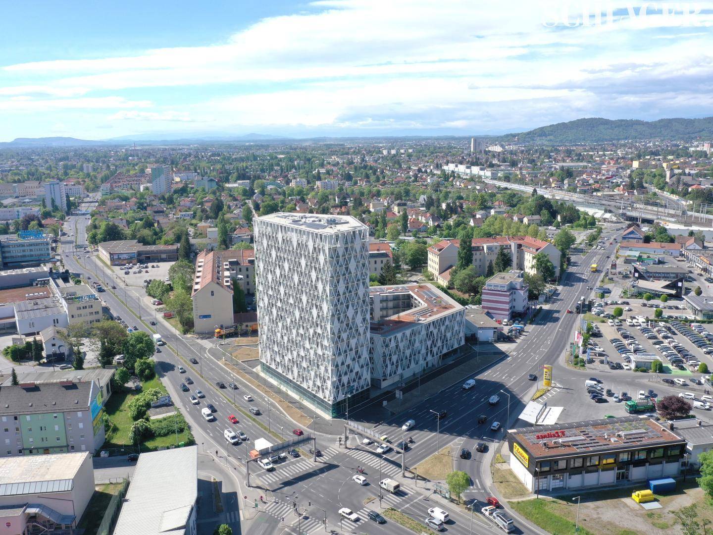 Weitläufige Luftaufnahme des modernen Gebäudes an einer belebten Kreuzung in städtischer Umgebung mit weitem Blick.