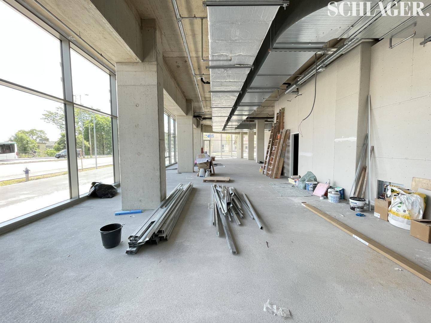 Unfertiger Gewerberaum mit großen Fenstern, Betonboden und sichtbaren Installationen an der Decke, bereit für den Ausbau.