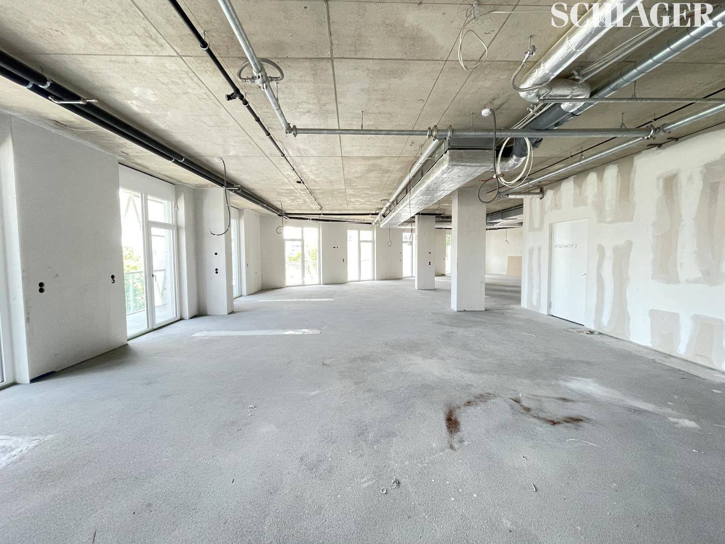 Großer, unfertiger Raum mit Betonboden, unverputzten Wänden und vielen Fenster-Tür-Elementen für viel Licht.