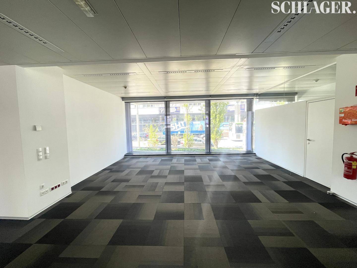 Heller Büroraum mit gemustertem Teppichboden und breiter Fensterfront zur Straße.
