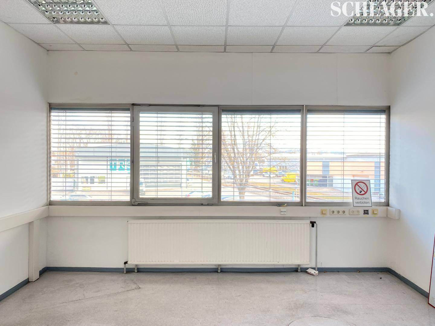 Leeres Bürozimmer mit großen Fenstern, Jalousien und einem Heizkörper unter der Fensterbank.