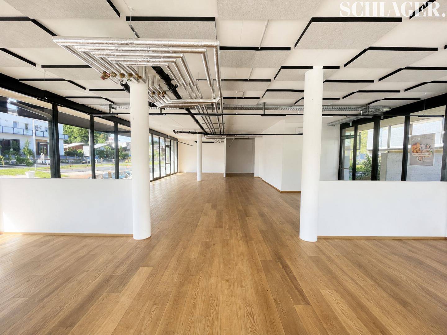 Geräumiger, offener Gewerberaum mit Säulen, großen Fenstern und sichtbaren Deckenleitungen.