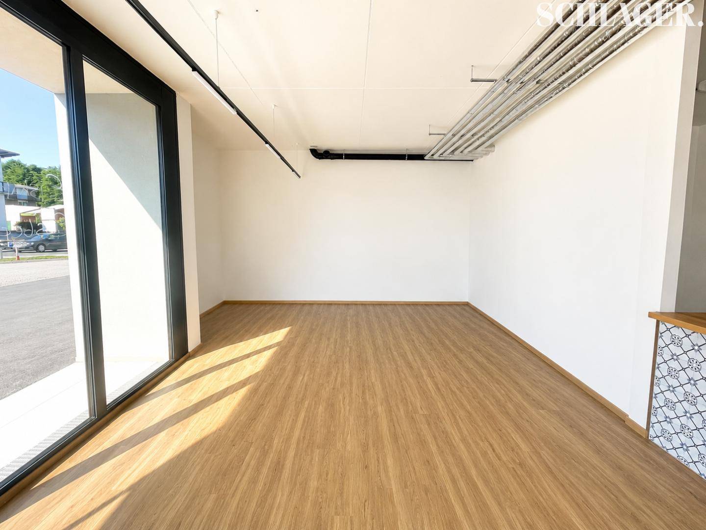 Heller, leerer Gewerberaum mit großen Fenstern, Holzboden und sichtbaren Deckeninstallationen.