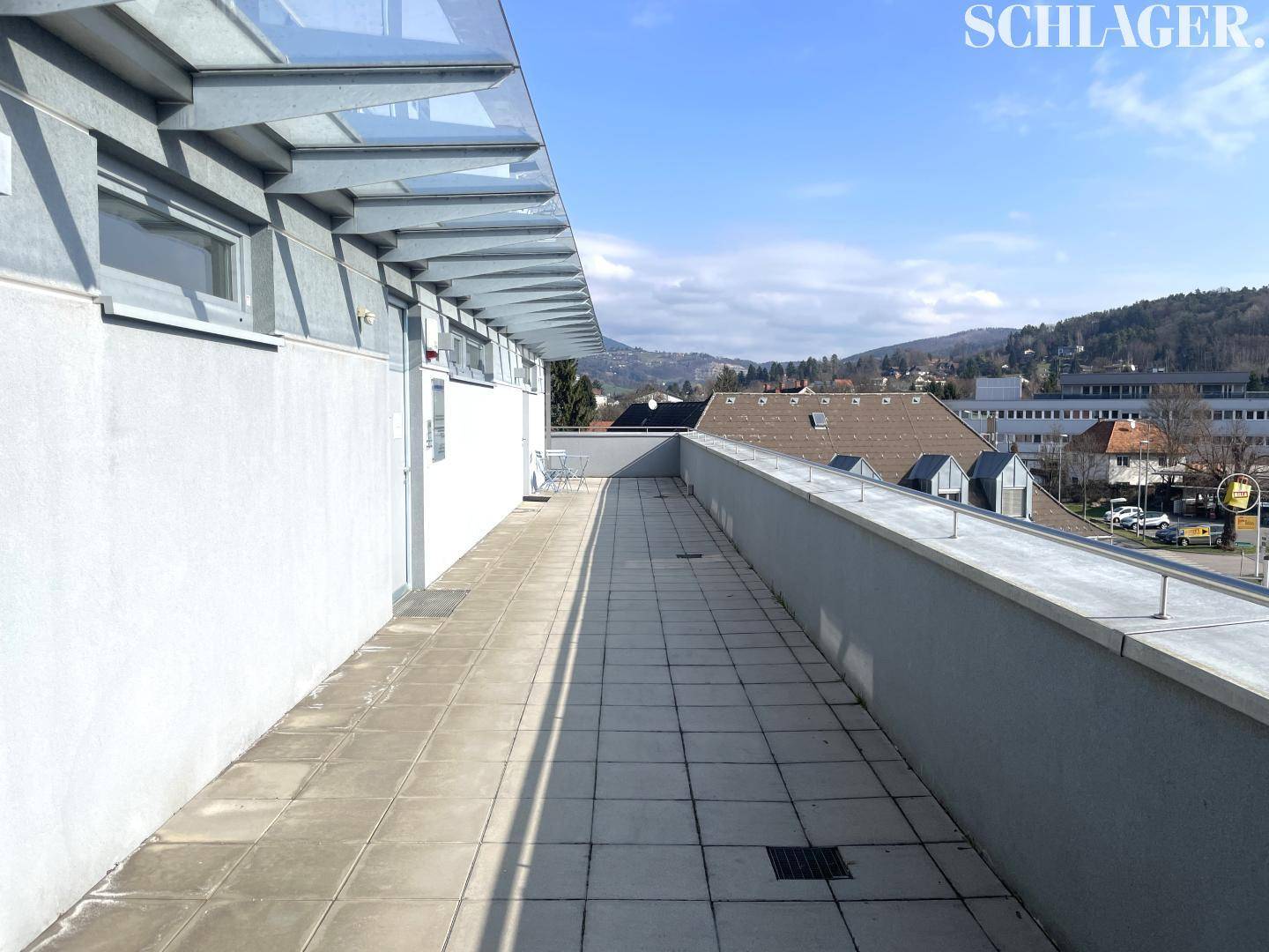 Lange, geflieste Terrasse mit Glasüberdachung und weitem Blick auf die umliegende Landschaft und Berge.