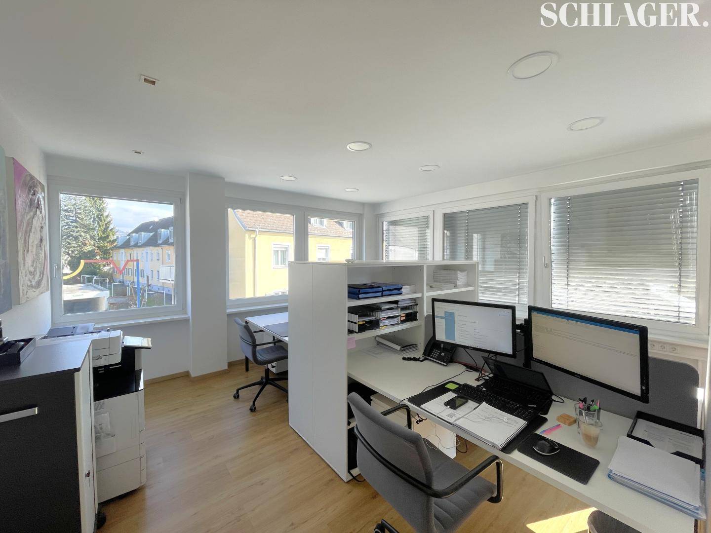 Geräumiges Büro mit mehreren Arbeitsplätzen, viel Tageslicht und Blick auf einen Spielplatz durch die Fenster.