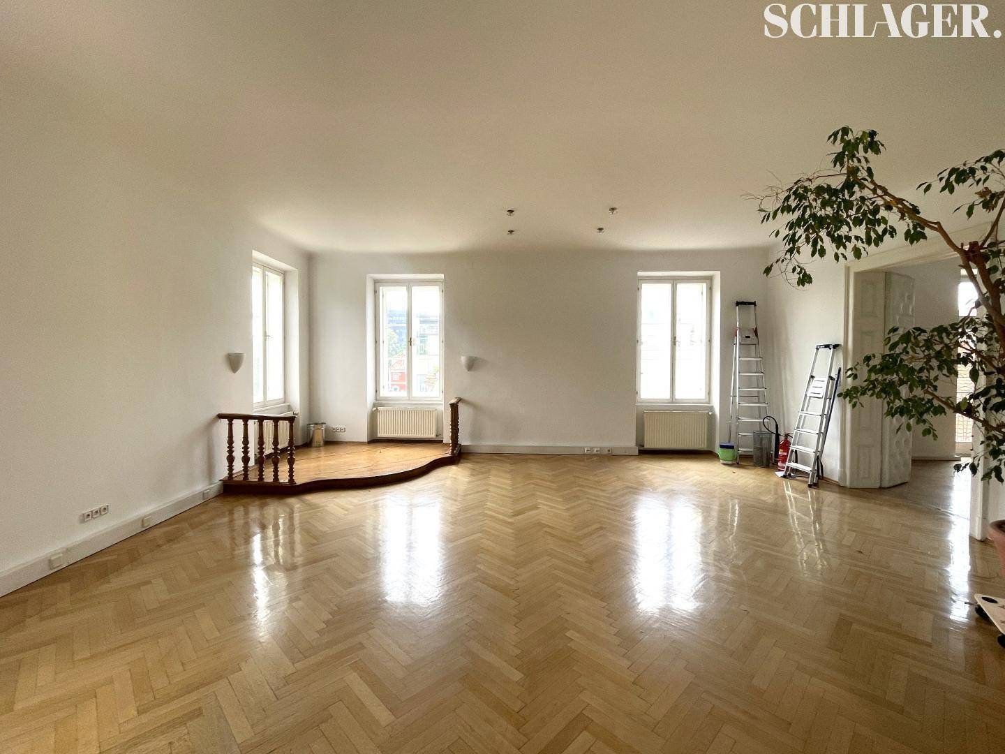 Geräumiges Wohnzimmer mit Fischgrätparkett, hohen Decken und viel natürlichem Licht durch große Fenster.