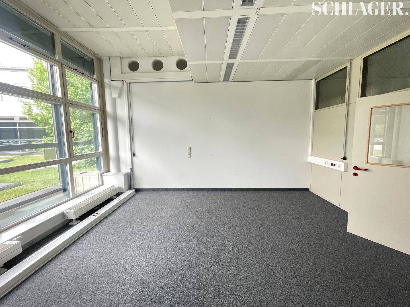 Geräumiger Büroraum mit großen Fenstern an der Seite, Teppichboden und einer schlichten Einrichtung.