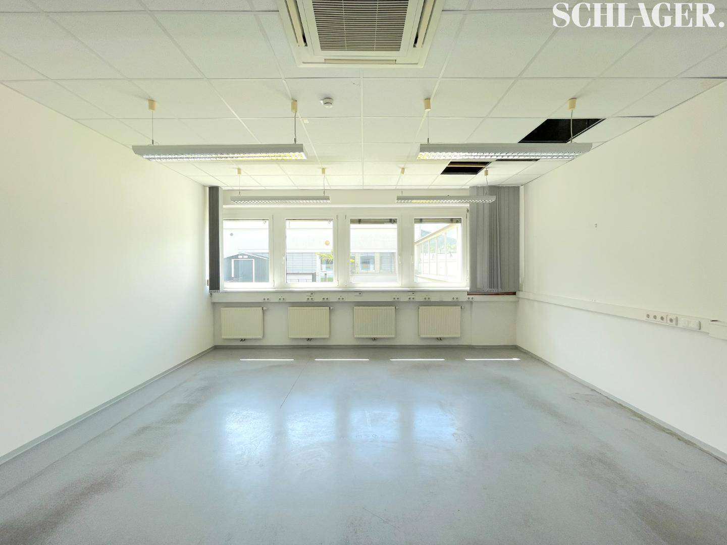 Heller, leerer Büroraum mit großen Fenstern, abgehängter Decke und Heizkörpern unter den Fenstern.