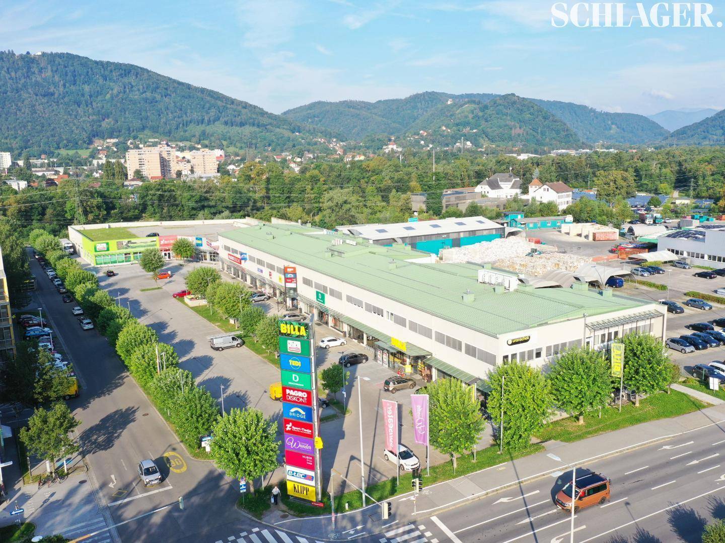 Luftaufnahme eines großen Geschäftskomplexes mit Parkplätzen, umgeben von Grünflächen und Bergen im Hintergrund.