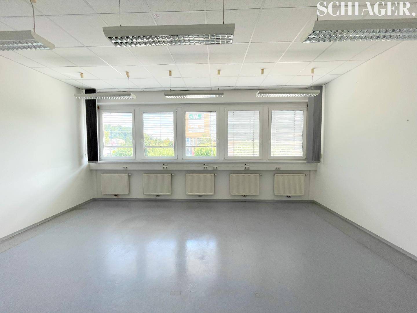 Geräumiger, unmöblierter Büroraum mit mehreren Fenstern, Jalousien und abgehängter Decke.