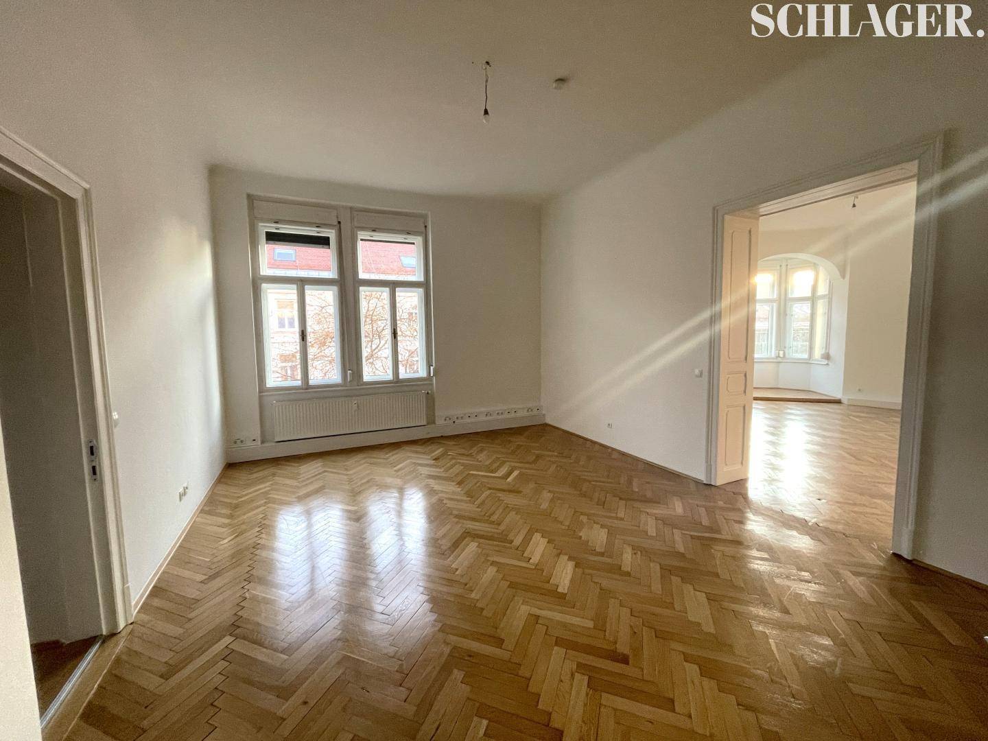 Geräumiges Zimmer mit glänzendem Fischgrätparkett und einem Durchgang zu einem weiteren hellen Raum.