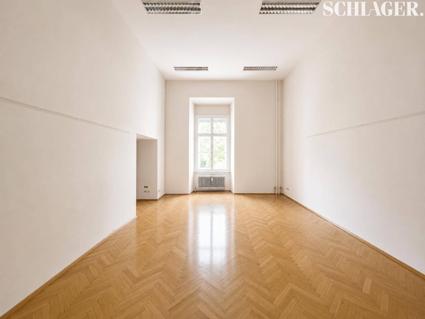 Geräumiger, heller Raum mit Fischgrätparkett, großem Fenster und schlichten Wänden.