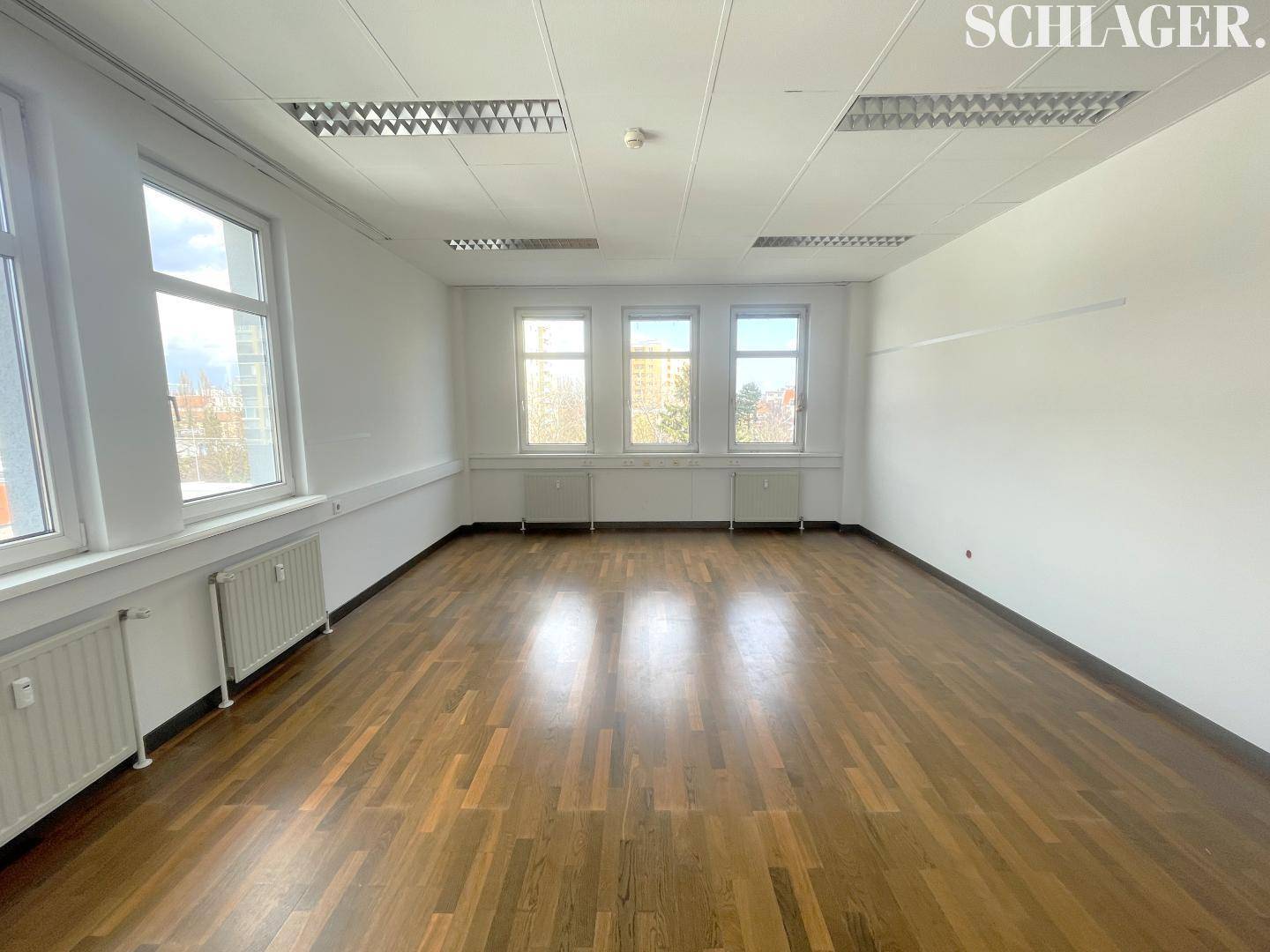 Leerer Büroraum mit dunklem Holzboden, weißen Wänden und einer Reihe von Fenstern für gute Belichtung.