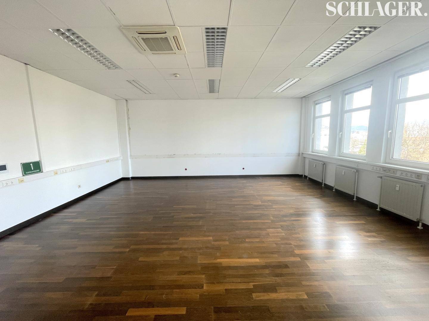 Geräumiger, unmöblierter Büroraum mit dunklem Holzboden, Klimaanlage und vielen Fenstern für Tageslicht.