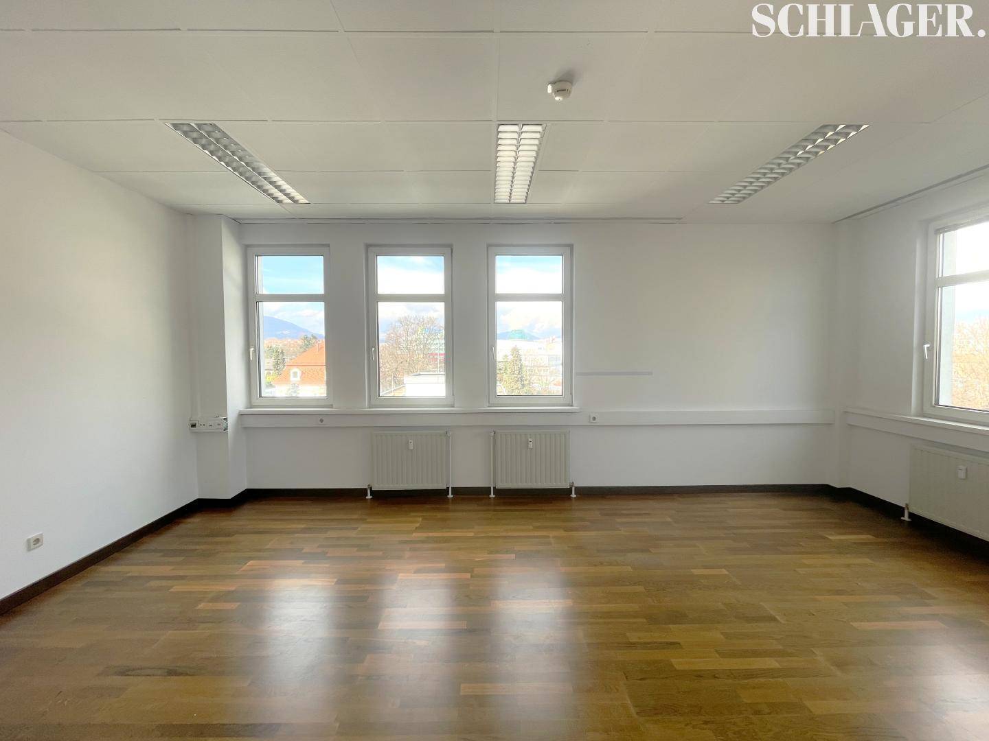 Heller Büroraum mit Holzboden und mehreren Fenstern, die viel natürliches Licht hereinlassen.