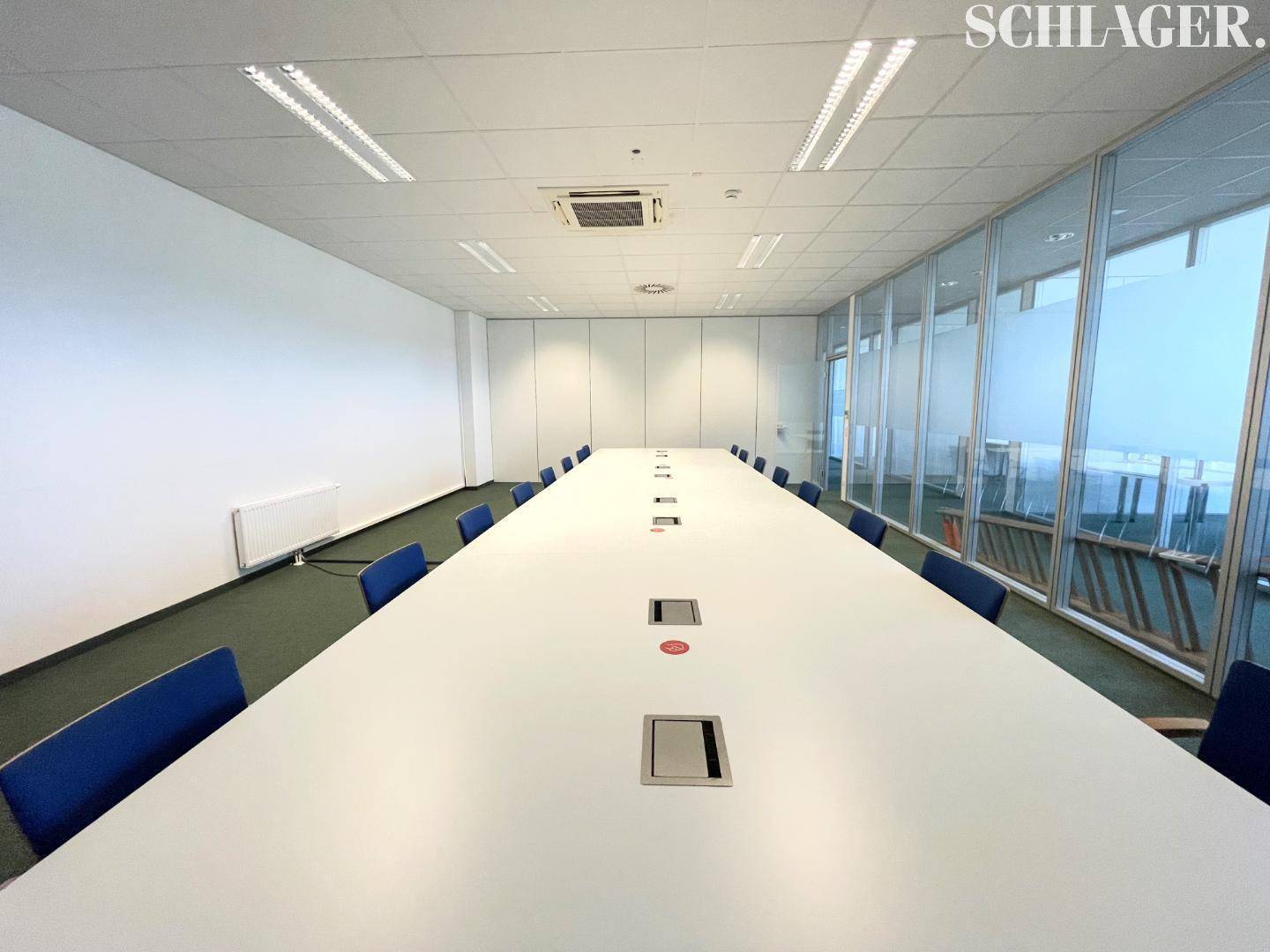 Moderner Konferenzraum mit langem weißen Tisch, blauen Stühlen und Glaswand für Besprechungen.
