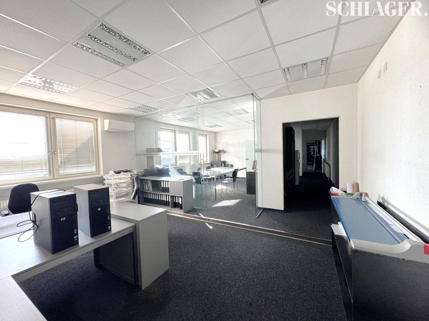 Helles Büro mit Teppichboden, großen Fenstern und Glastrennwand zu einem weiteren Arbeitsbereich.