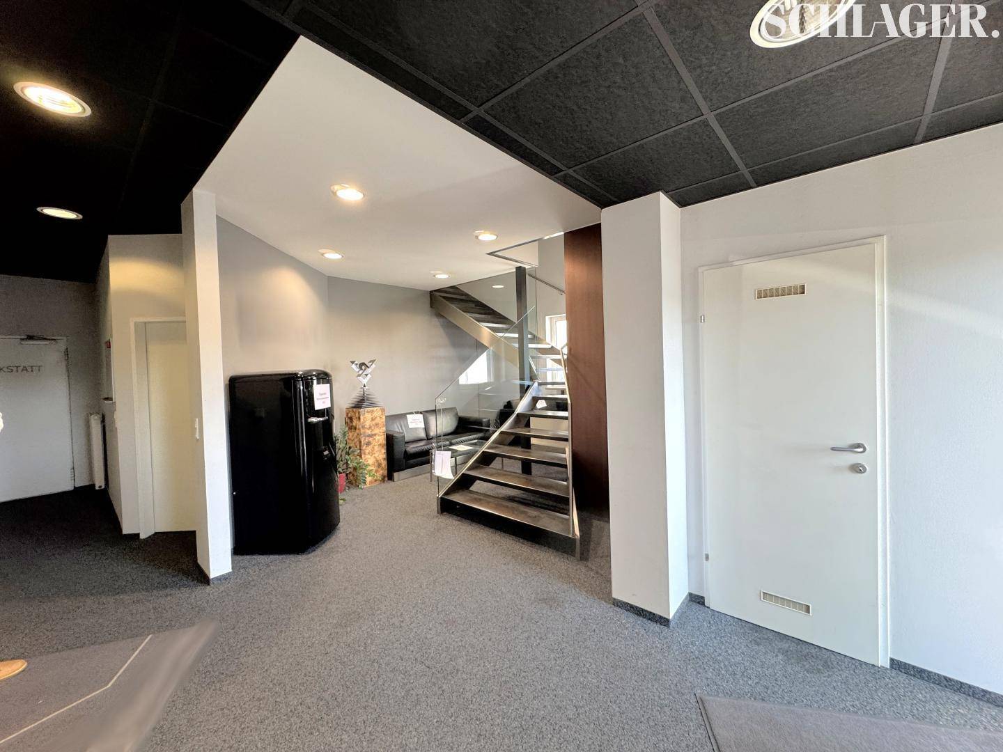 Moderner Empfangsbereich mit Teppichboden, offener Treppe mit Glasgeländer und dunkler Decke.