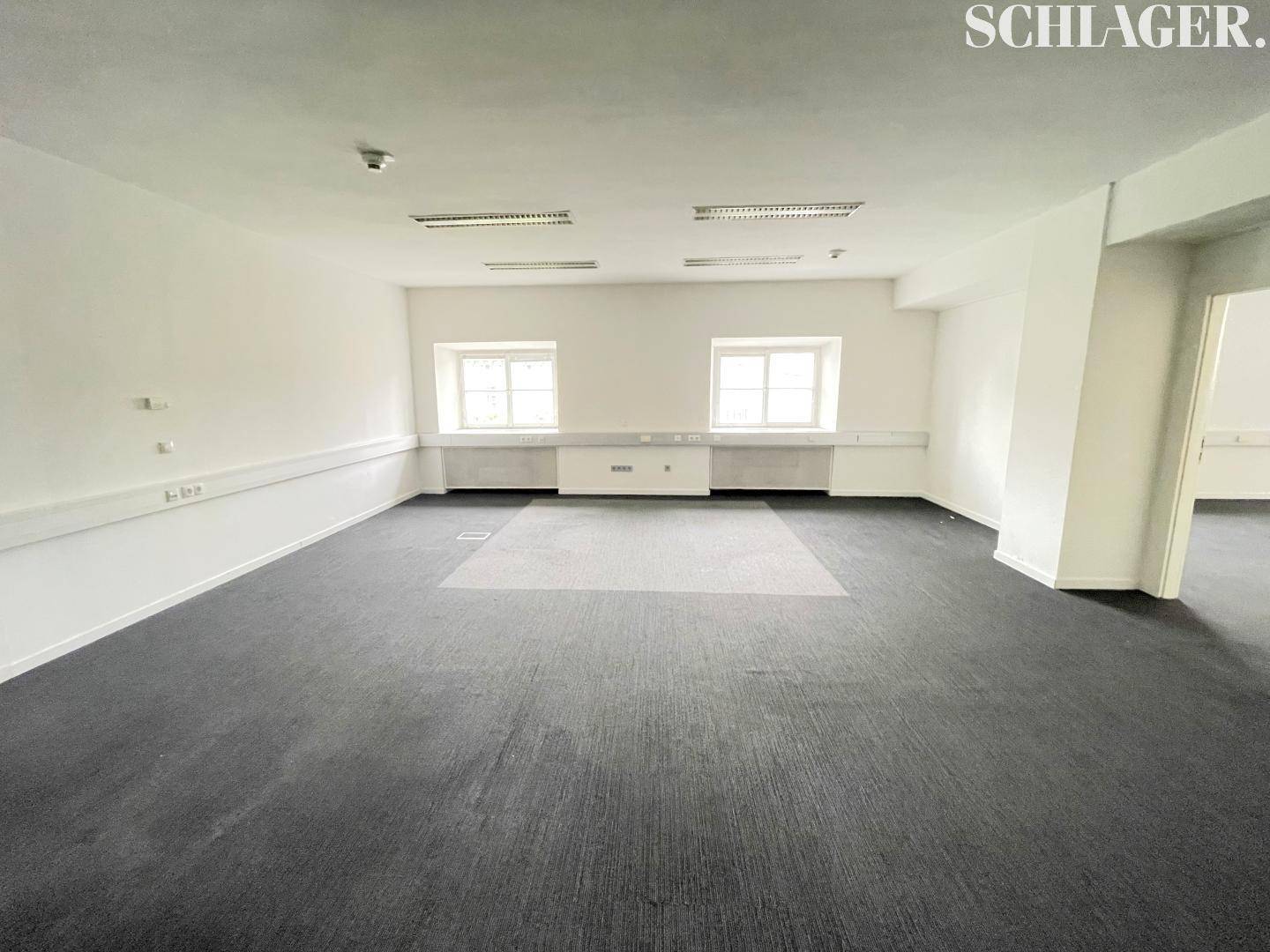 Großer, leerer Büroraum mit dunklem Teppichboden und zwei Fenstern für Tageslicht.