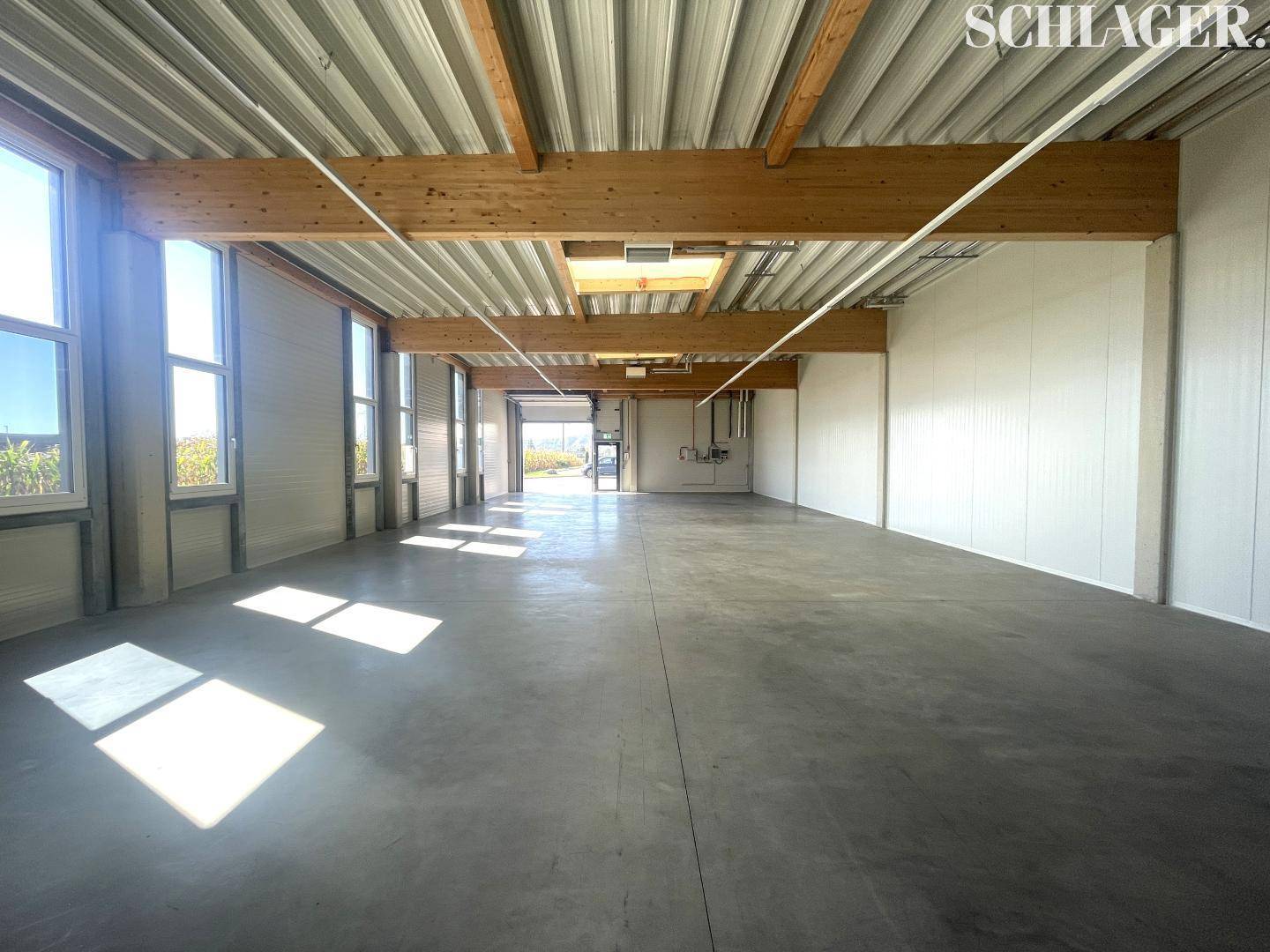 Helle Lagerhalle mit Betonboden, Holzbalken und einem großen Rolltor, das den Blick nach draußen freigibt.