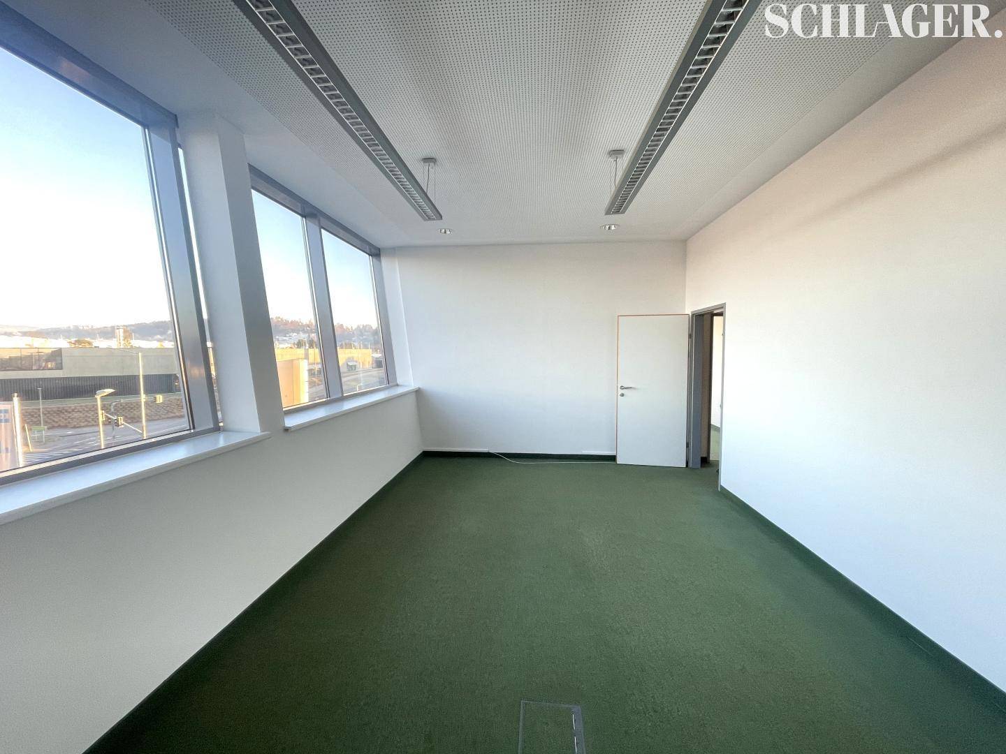 Helles, leeres Büro mit großen Fenstern, grünem Teppichboden und weiß gestrichenen Wänden.