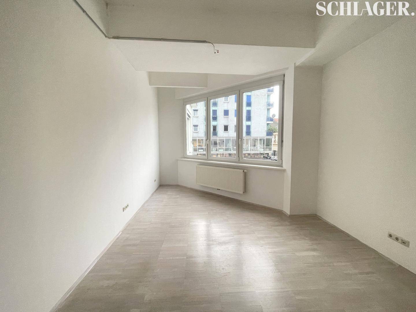 Leerer Raum mit hellen Holzböden, weißen Wänden und einem Erkerfenster, das einen Blick auf umliegende Gebäude bietet.