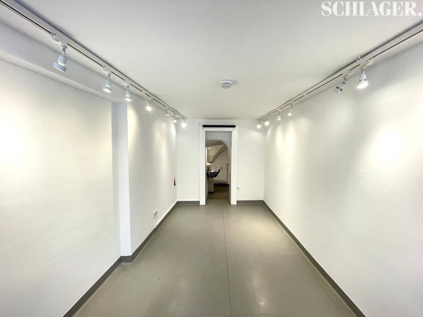 Langer, schmaler Verkaufsraum mit weißen Wänden, Schienenspots an der Decke und grauem Boden.