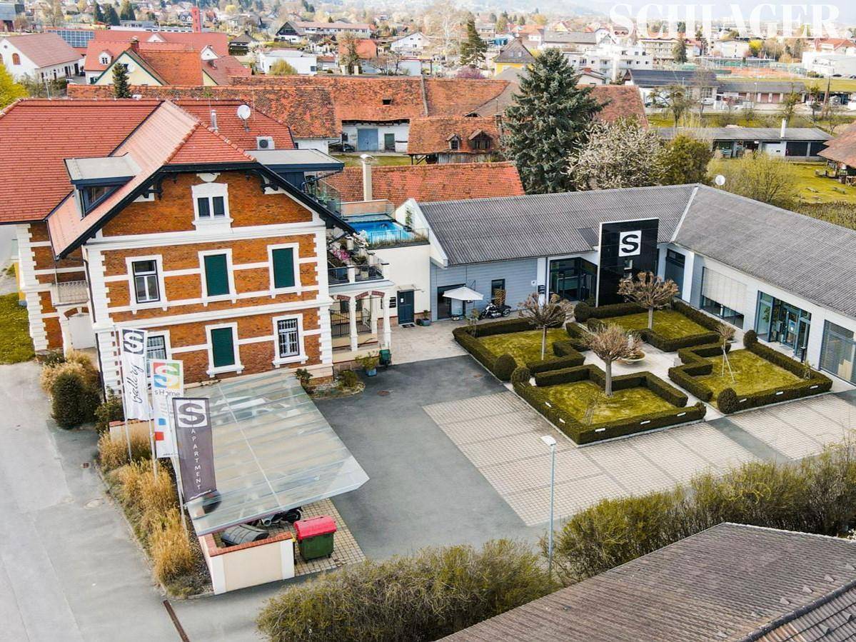 Luftaufnahme eines Geschäftskomplexes mit historischem Gebäude, modernen Anbauten, Parkplätzen und gepflegten Grünflächen.
