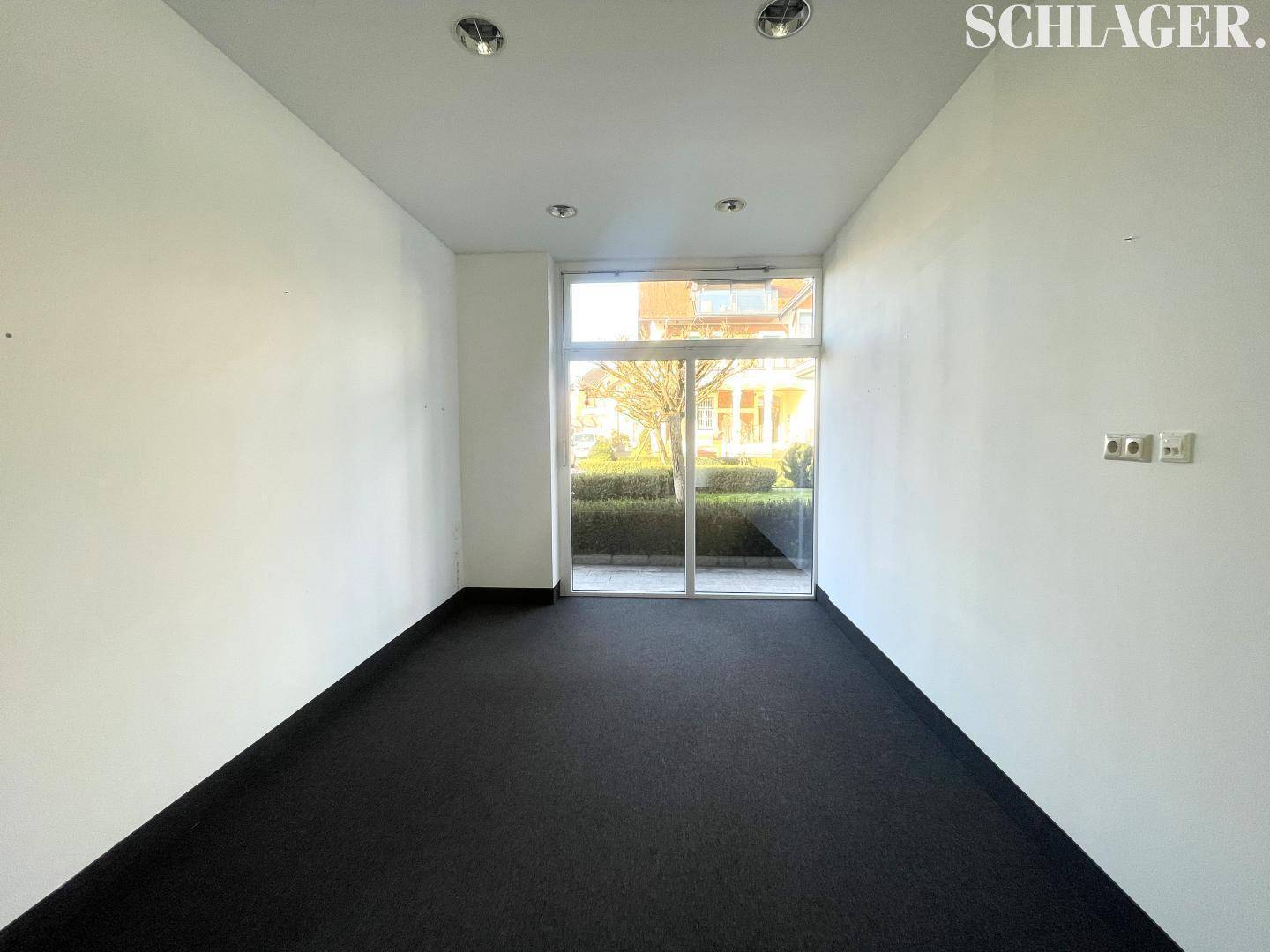 Leerer Geschäftsraum mit dunklem Teppichboden und großer Glasfront, die viel Tageslicht hereinlässt.